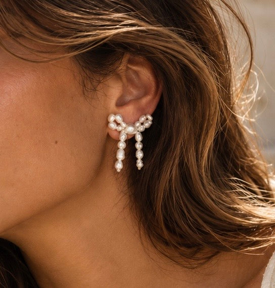 A close-up of a model's ear wearing a dangling pearl bow earring with small pearl drops hanging from the bow.